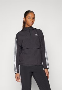 Svart atletisk jakke med kvart glidelås, hette og lommer foran. Har hvite adidas-striper langs ermene. Lett materiale.
