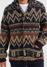 Hombre vistiendo un cárdigan de punto abotonado con patrones geométricos y zigzagueantes en azul marino, beige, rojo y verde, con las manos en los bolsillos.