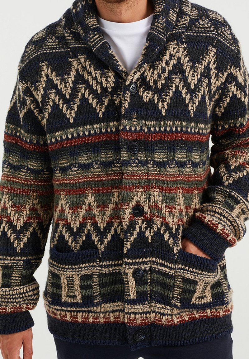 Hombre vistiendo un cárdigan de punto abotonado con patrones geométricos y zigzagueantes en azul marino, beige, rojo y verde, con las manos en los bolsillos.