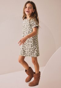 Jeune fille souriante, portant une robe à manches courtes à imprimé léopard avec poche avant et bottes en daim marron, debout devant un fond neutre.