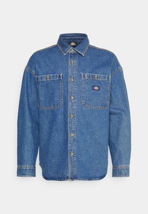 Camicia di jeans blu con maniche lunghe, chiusura con bottoni sul davanti e due tasche anteriori. Presenta cuciture gialle e un piccolo logo sulla tasca sinistra.