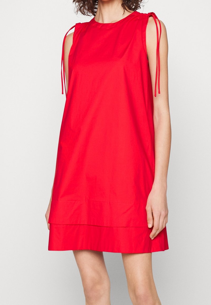 Robe rouge vif sans manches avec des bretelles nouées aux épaules et un ourlet ample et superposé portée par une personne aux cheveux bouclés sur un fond uni.