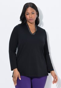Vrouw in een zwart longsleeve shirt met een kralen halslijn en paarse broek, staande voor een witte achtergrond.