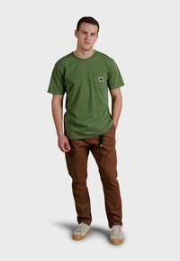 Olivově zelené bavlněné tričko s kapsou, kombinované s hnědými kalhotami a krémovými teniskami s gumovou podrážkou a tkaničkami. Jednoduchý design, uvolněný střih.