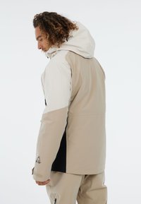 Beige en witte waterdichte jas met een capuchon, ritsen en contrasterend zwart zijpaneel. Heeft een soepele textuur en verstelbare manchetten.
