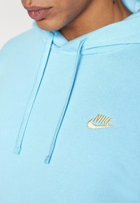 Acercamiento de una persona que lleva puesta una sudadera Nike de color azul claro con un logo bordado en dorado y cordones, mostrando parte de la cara y el cuello.