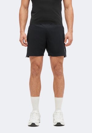 Mann trägt schwarze Sportshorts, schwarzes T-Shirt, weiße Sportsocken und weiße Turnschuhe, steht mit entspannten Armen an den Seiten.