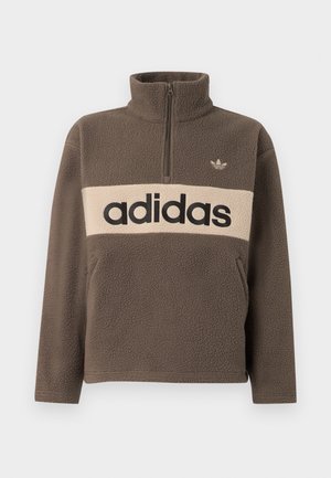 Homme portant un pull en polaire marron avec une bande horizontale beige et un grand logo noir "adidas", debout devant un fond blanc uni.