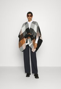 Poncho multicolor con un patrón geométrico en gris, negro, naranja y blanco, con un cinturón de lazo y una tela suave y texturada.