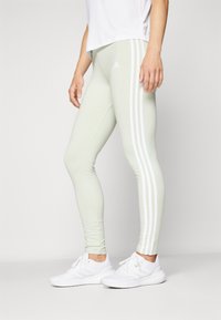 Svetlo zelene legginge iz raztegljivega materiala, s belimi stranskimi črtami in logotipom Adidas na zgornjem levem stegnu.