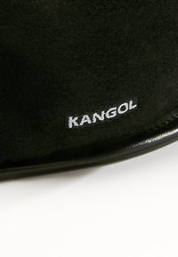 Svart texturerad tyg med vitt broderat "KANGOL"-logotyp på en böjd yta, sannolikt del av en hatt eller klädesplagg.