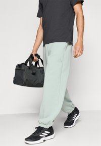 Ανοιχτό πράσινο φόρμα με ελαστικά μανσέτα, σκούρο γκρι μπλουζάκι και μαύρα αθλητικά παπούτσια με λευκές ρίγες. Κρατάει μια σκούρα αθλητική τσάντα.