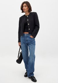 Giacca cropped nera con texture e bottoni dorati, abbinata a pantaloni in denim a gamba larga blu chiaro e una borsa nera, indossata sopra a una maglietta nera.