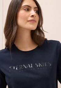 Camiseta de manga larga azul marino oscuro con texto metálico plateado "CIELOS ETERNOS" y "ESPEJO DE PENSAMIENTOS" en la parte frontal, material de algodón suave.