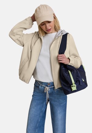 Frau mit beiger Mütze, cremefarbener Jacke mit Reißverschluss, weißem Hemd, blauer Jeans, trägt eine marineblaue Tasche mit grünen Reißverschlussanhängern auf der Schulter.