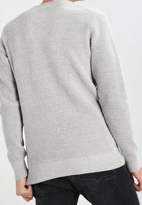 Person klädd i en ljusgrå texturerad stickad tröja och svarta jeans, sedd bakifrån med händerna nära höfterna mot en enfärgad bakgrund.