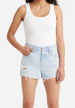 Femme portant un débardeur blanc côtelé sans manches et un short en jean bleu clair déchiré avec des ourlets effilochés, une main dans la poche.