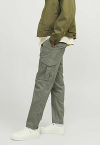 Pantalones cargo de algodón gris claro con bolsillos laterales, combinados con una camisa beige y una chaqueta verde. Zapatillas blancas con acentos texturizados.