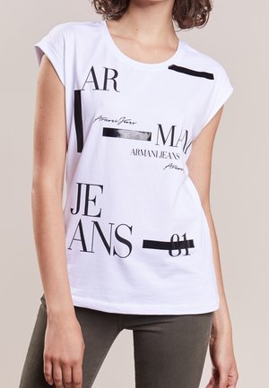 Femme portant un top blanc sans manches avec un texte abstrait noir et des graphismes indiquant "Armani Jeans", associé à un pantalon foncé.
