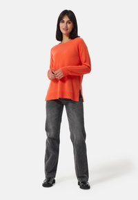 Maglione arancione a maglia con maniche lunghe, scollo rotondo e spacchi laterali abbinato a jeans larghi grigio scuro e scarpe nere.