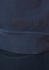 Felpa blu con orlo a coste, presenta una stampa scura con "L.P." e un design a chevron. Il materiale è morbido e la texture appare liscia.
