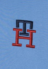 Tela azul claro con un logo bordado en rojo y azul oscuro que presenta las letras entrelazadas "T" y "H" y una "H" destacada en el centro.