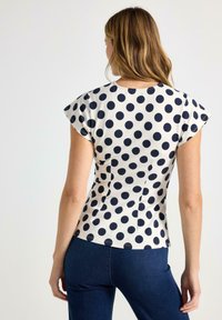 Kortærmet bluse i cremefarvet stof med marineblå polkaprikker, tætsiddende design og detaljer med bageste søm. Kompletteret med mørke denimbukser.