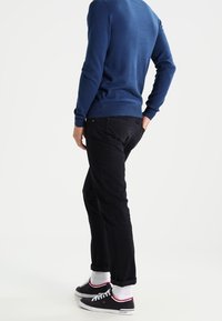 Blauer Pullover mit gerippten Bündchen, kombiniert mit schwarzen Hosen. Schwarze Sneakers mit weißen Sohlen und roten Akzenten, getragen mit weißen Socken.
