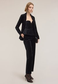 Kobieta w czarnym aksamitnym żakiecie i bezrękawnikowej bluzce z pasującymi spodniami oraz przezroczystymi skarpetkami w panterkę i czarnych sandałach na wysokim obcasie.