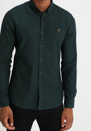 Homme portant une chemise boutonnée à manches longues vert foncé avec un petit logo jaune sur la poitrine gauche, associée à un pantalon noir.