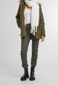 Olivově zelená parka s kožešinovým lemováním, bílá šála, přiléhavý bílý top, úzké olivové kalhoty a černé robustní boty. Hladké textury a vrstvený design.