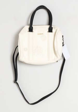 Sac à main crème avec poignées noires et longue bandoulière, motifs floraux en relief, logo de marque doré et une étiquette blanche attachée sur le côté.