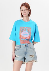 Μπλε oversized μπλουζάκι με πολύχρωμο γραφικό σχέδιο που απεικονίζει χείλη και κείμενο, σε συνδυασμό με φθαρμένα τζιν σορτσάκια. Μαλακό βαμβακερό ύφασμα, χαλαρή εφαρμογή.