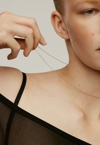 Collar de cadena de oro sostenido entre los dedos, con una textura delicada. La modelo lleva una blusa negra transparente con tirantes finos delante de un fondo neutro.