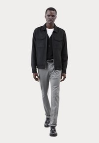 Giacca nera con due tasche frontali sopra un cardigan nero, t-shirt bianca, pantaloni grigi e scarpe nere; texture liscia in tutto.