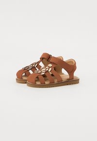 Brune skinnsandaler med åpen design, med tre stropper, borrelås og en skilpadde-formet dekorasjon foran. Flat såle.