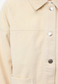 Beige corduroy jakke med lodret ribbing, krave og sølv trykknapper; inkluderer en lomme foran.