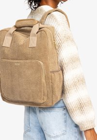 Zaino beige testurizzato con un motivo a righe, doppi manici, tasca laterale con cerniera e spallacci regolabili. Indossato sopra un maglione chiaro.
