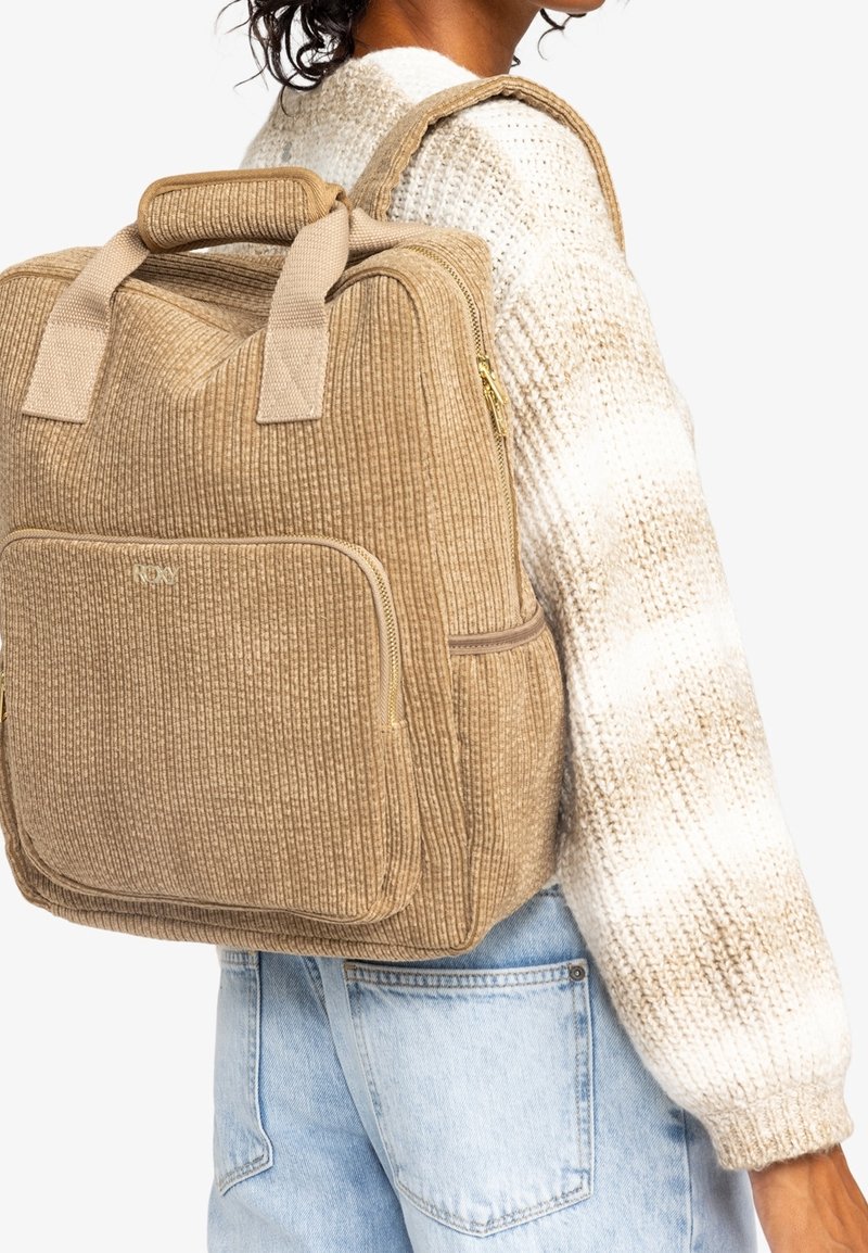Zaino beige testurizzato con un motivo a righe, doppi manici, tasca laterale con cerniera e spallacci regolabili. Indossato sopra un maglione chiaro.
