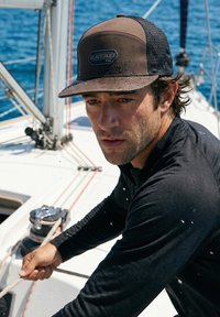 Hombre con gorra y camisa negras tirando de una cuerda en un velero con agua azul de fondo, gotas de agua en su cara y ropa.