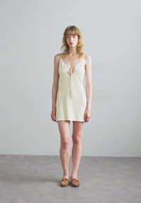 Cremfarbenes, ärmelloses Minikleid mit V-Ausschnitt und Bindebindung vorne, glatte Textur und gerader, taillierter Schnitt. Kombiniert mit braunen Schuhen.