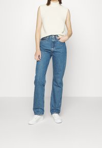 Light beige sleeveless top and high-waisted blue denim jeans, featuring a straight-leg cut, with white sneakers. Simple, clean design.