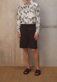 Camicia a maniche lunghe con bottoni, caratterizzata da un motivo astratto bianco e nero, abbinata a pantaloni corti neri e semplici sandali neri a infradito.