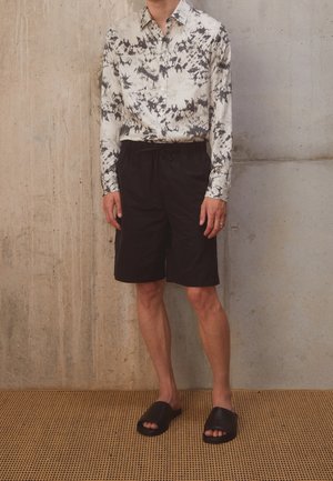 Lange mouwen, knoopjeshemd met een zwart-wit abstract patroon, gecombineerd met zwarte shorts en eenvoudige zwarte slippers.