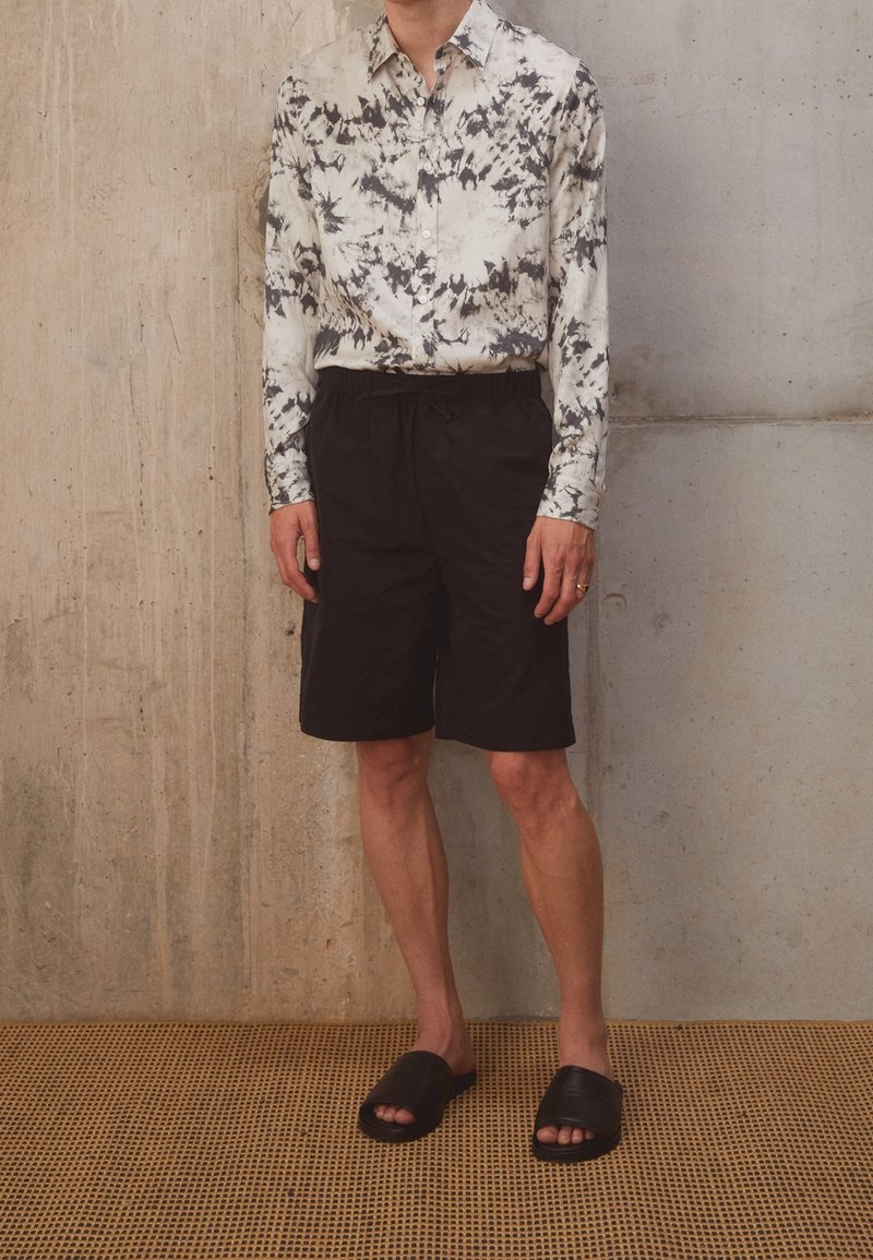 Camicia a maniche lunghe con bottoni, caratterizzata da un motivo astratto bianco e nero, abbinata a pantaloni corti neri e semplici sandali neri a infradito.