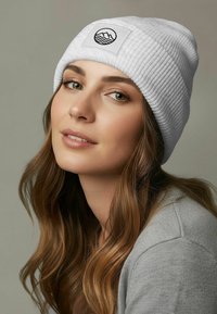 White ribbed knit beanie with a logo patch featuring waves. The beanie is soft textured, with a snug fit, worn slightly rolled at the edge.