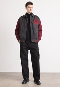 Gray and burgundy varsity jacket with embroidered logo, snap buttons, ribbed cuffs, paired with black cargo pants and black shoes.