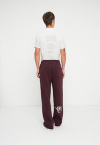 Hvid kortærmet skjorte med et mesh-panel på ryggen, parret med bordeaux-farvede sweatpants med et gult og hvidt grafisk design.