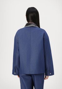 Blue denim jacket with a black leather collar, oversized shape, long sleeves, and side slits. Stitching details are visible along the seams.