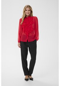 Frau mit langen blonden Haaren, die eine transparente rote Bluse mit Rüschen, schwarze Hose und schwarze spitze High Heels trägt, vor einem schlichten Hintergrund steht.